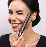 Una donna sorride ampiamente mentre tiene un pennello per mascara, mostrando il suo entusiasmo per la bellezza e l'applicazione del trucco.