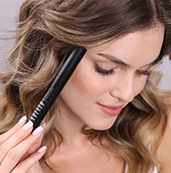 Una donna con capelli castani mossi tiene un tubetto di mascara nero vicino al viso, guardando in basso e sorridendo leggermente.